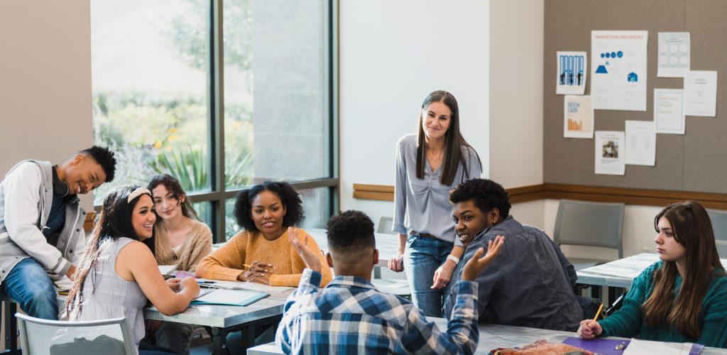 The diverse class of high school students and the mid adult female teacher listen to a classmate as he gestures and speaks.